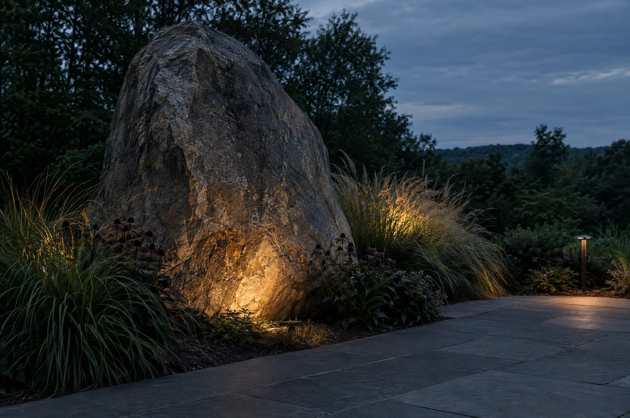 Illustration of the direction Curb's is heading: a massive boulder set into the ground beside large charcoal pavers and ornamental grasses, lit warmly at dusk