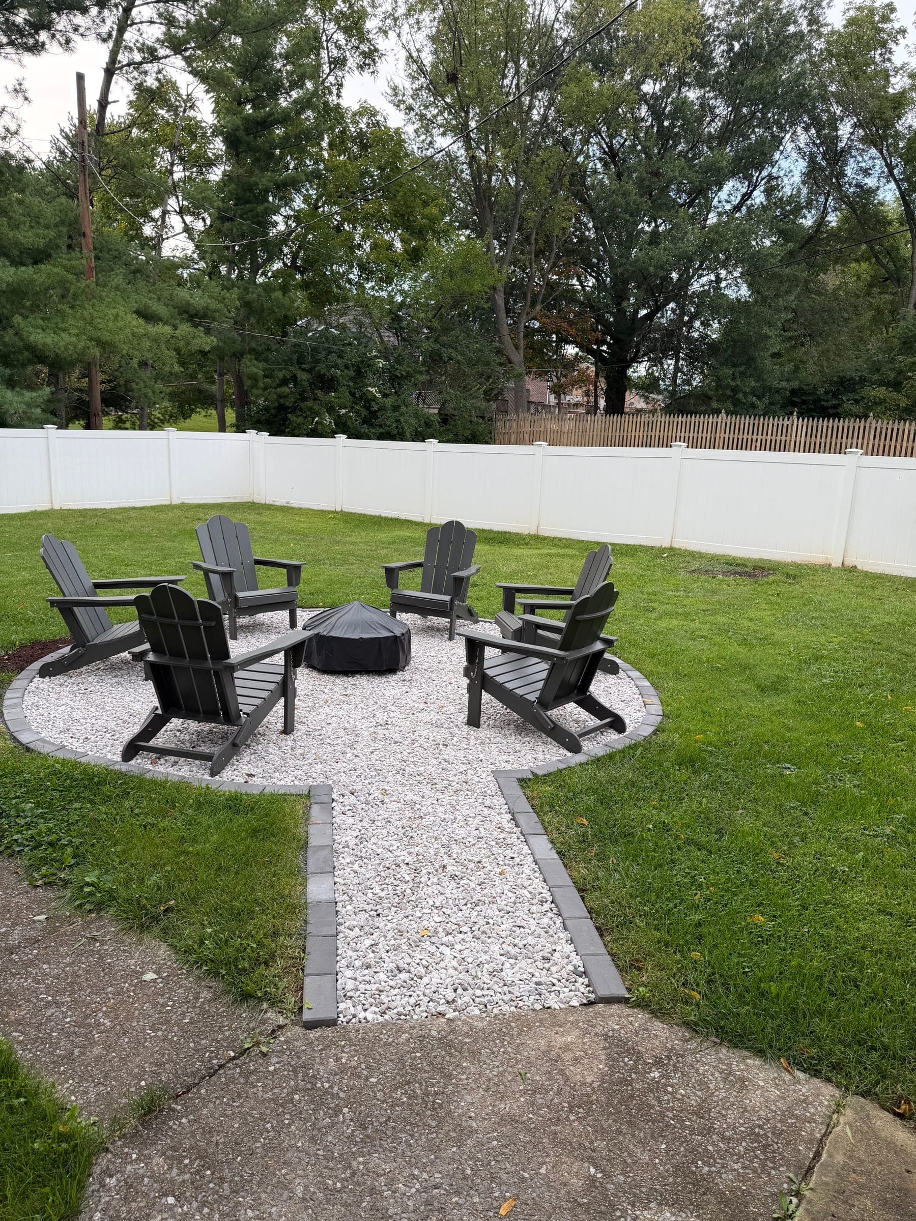 A finished backyard fire-pit patio: pale gravel edged with grey pavers, six dark Adirondack chairs, a mown lawn and a white fence behind